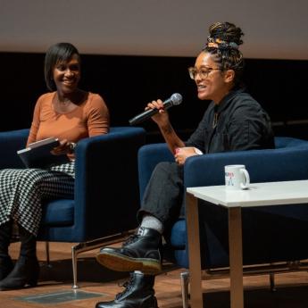 Eve L Ewing sits next to Sasha-Ann Simons on stage at an event. Eve is holding a microphone while talking.