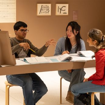Three students engaged in discussion while sitting at a table. Exhibit artwork is visible behind them.