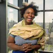 Gabrielle Randle-Bent wearing a lime green scarf laughs in front of windows. Outside the campus buildings are visible.