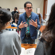 From behind two students Edgar Garcia is speaking and gesturing while standing in front of an open book.